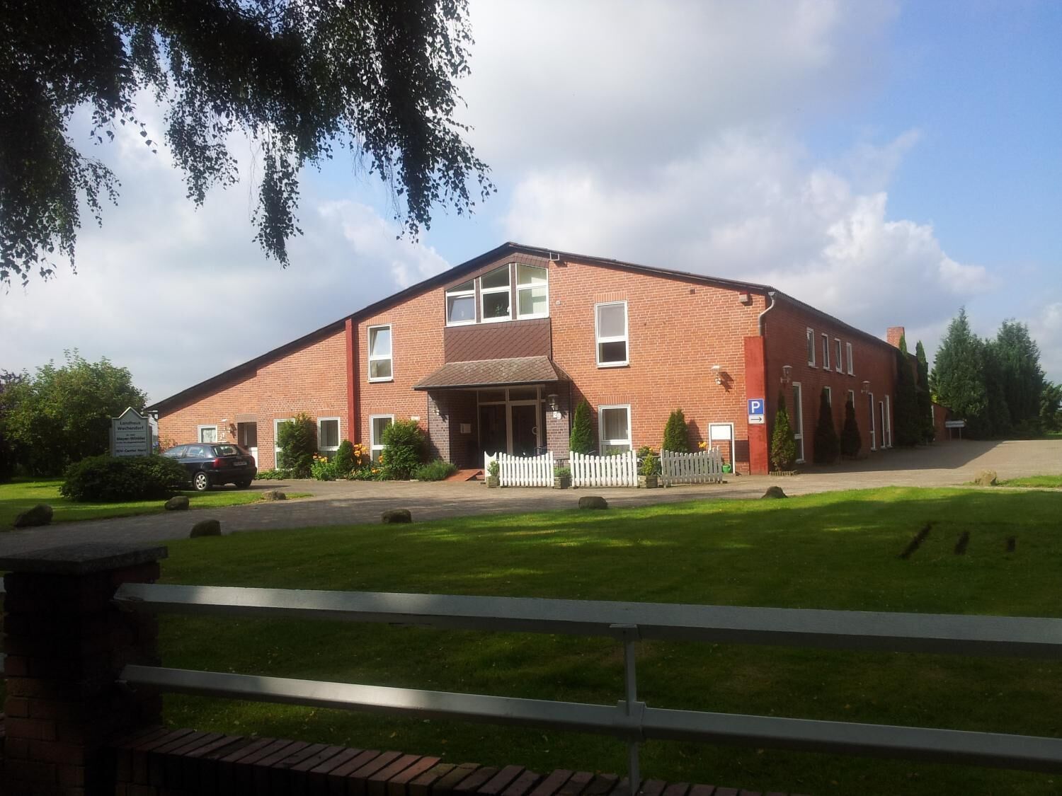Un bâtiment en briques de deux étages, doté d’une entrée centrale, d’une allée bordée d’une clôture et d’un panneau indiquant un espace de stationnement.