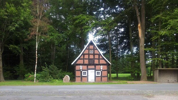A small, traditional building with a white door and windows, surrounded by trees. In front of the building, there is a paved area with two chairs and a table. - Hotel Landhaus Wachendorf (Bruchhausen-Vilsen)