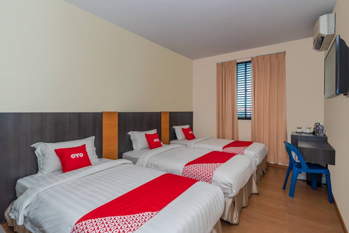 A hotel room with two beds, each with a red and white bedspread, a wooden headboard, and a red pillow with "OYO" branding. There is a desk with a chair, a television mounted on the wall, and a window with curtains.
