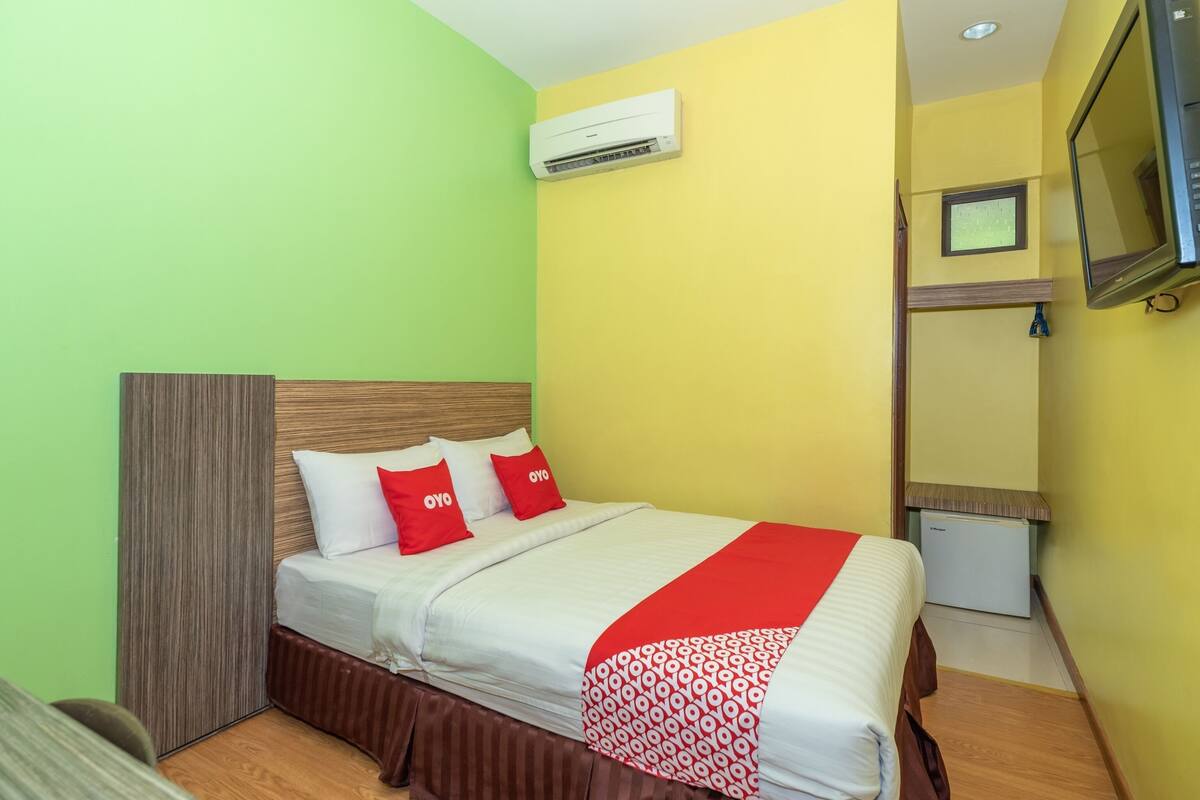 A hotel room with a bed, a TV, an air conditioner, and a small refrigerator.
