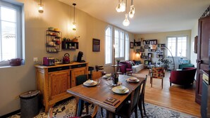 A spacious living area with a wooden dining table set for four, surrounded by comfortable seating. A kitchen area with wooden cabinets and a built-in oven is visible to the left. - l'En Vert (Bagnères-de-Bigorre)