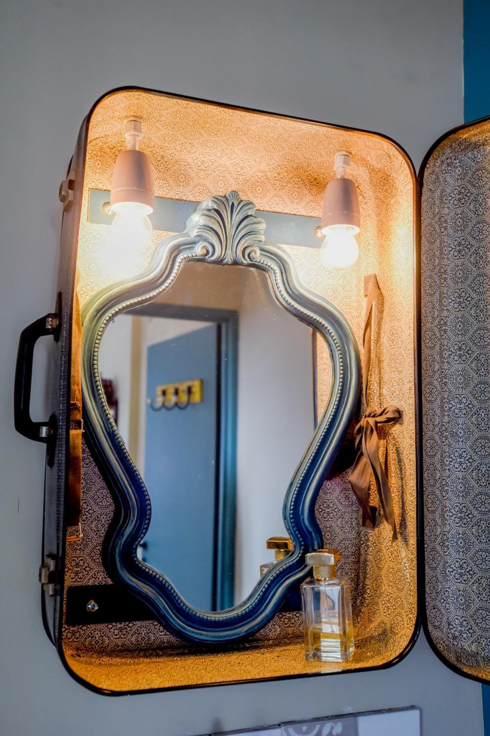 Un miroir de style vintage, doté d’un cadre décoratif et présenté dans un étui ressemblant à une valise, et équipé de deux lampes.