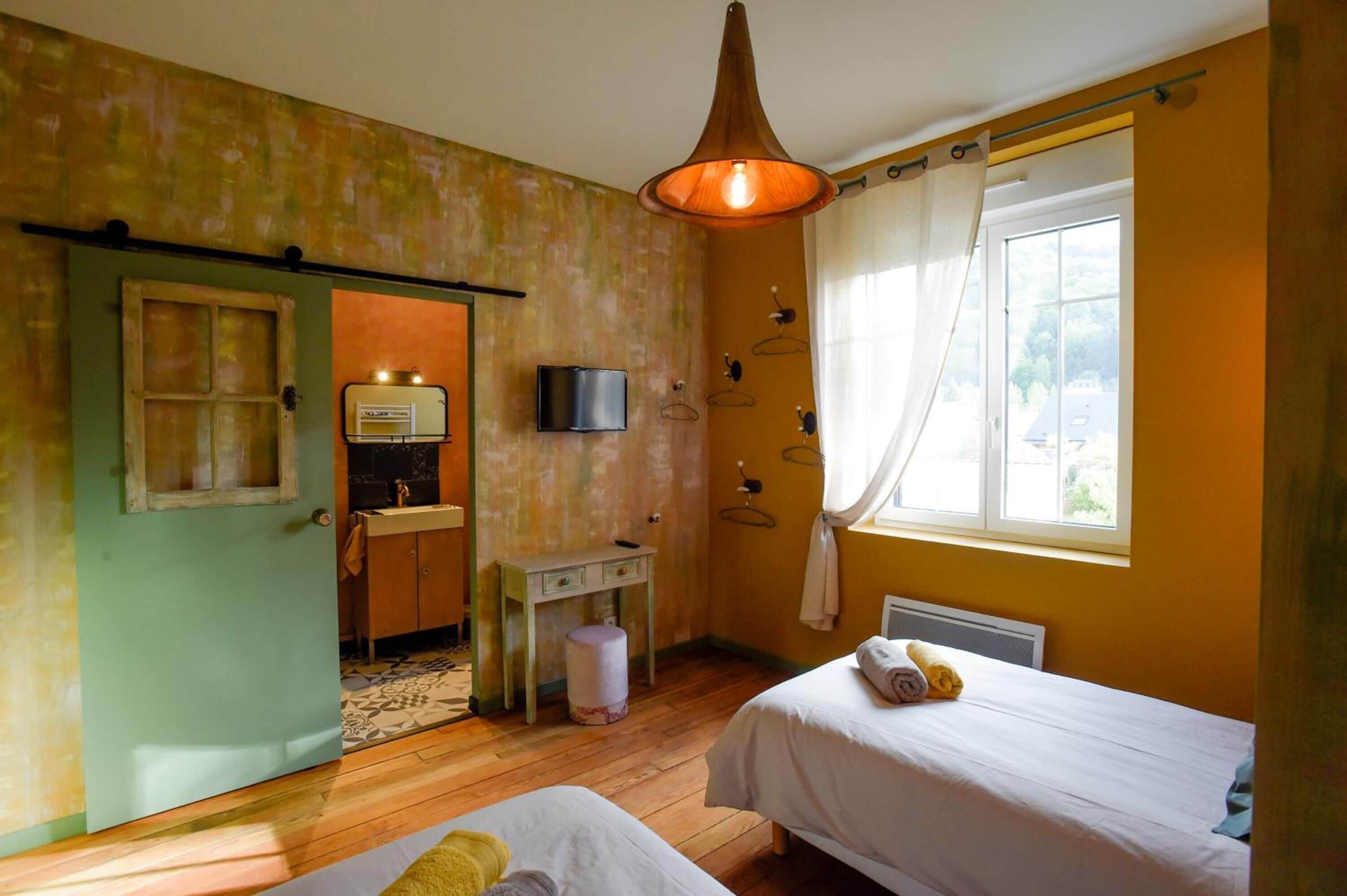 Een kamer met twee bedden, een houten vloer, een groene deur, een klein tafeltje met een lamp en een raam met gordijnen.
