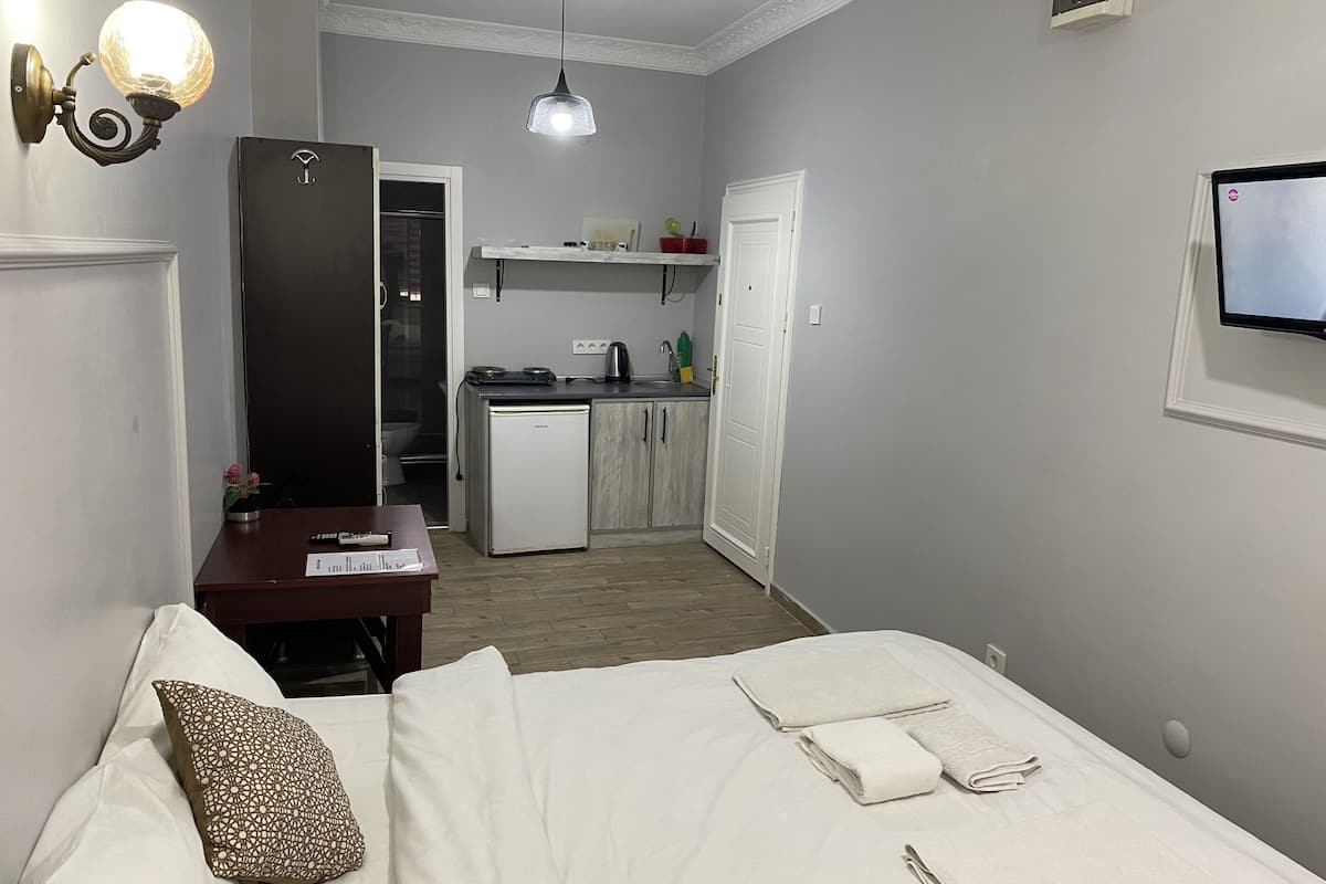 A bedroom with a bed, a desk, a small kitchenette, and a TV mounted on the wall.