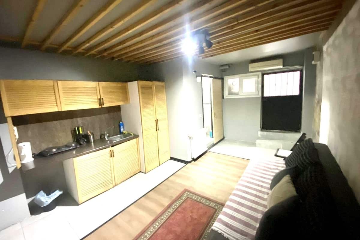 A compact kitchen with wooden cabinets, a sink, and a small countertop. There is a window with bars, a door, and a patterned rug on the floor.