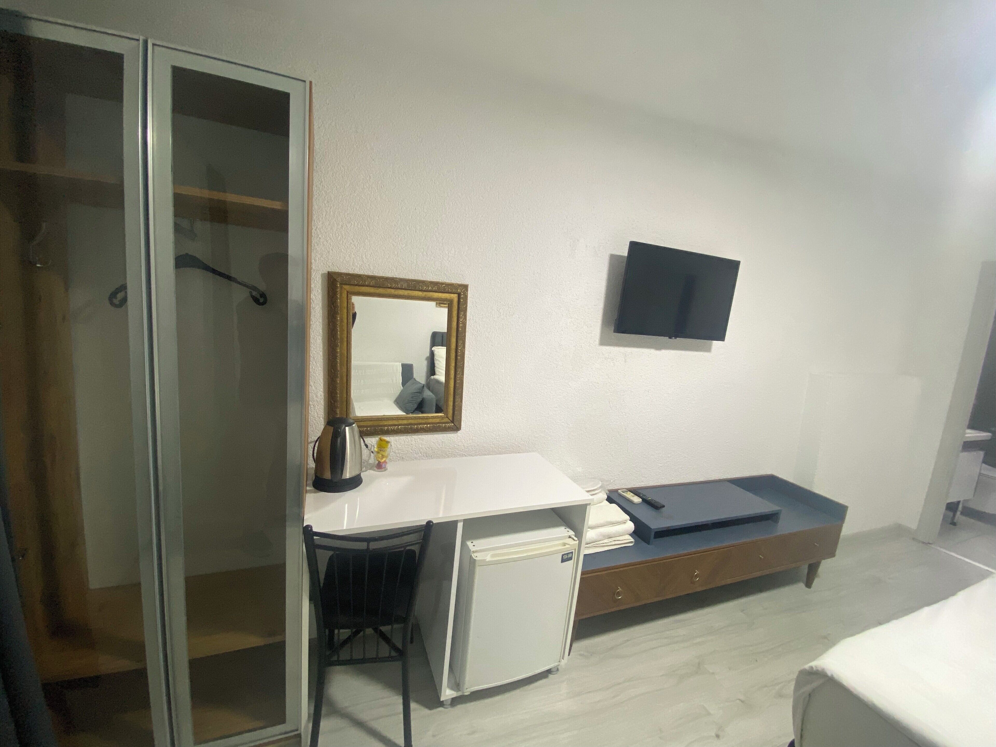 Une chambre d’hôtel dotée d’un placard avec une porte coulissante en verre, d’un petit bureau avec une chaise, d’un téléviseur à écran plat fixé au mur et d’un lit recouvert d’une couverture blanche.