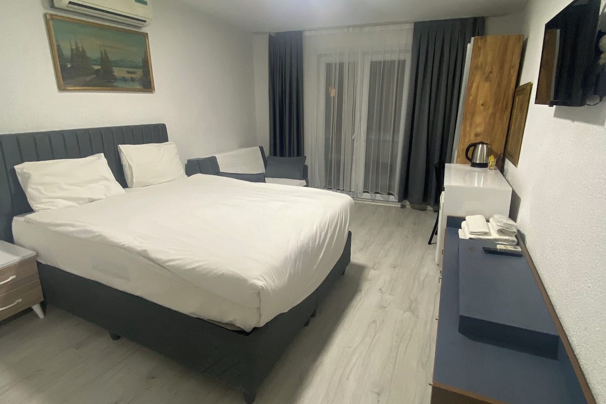 A modern hotel room with a large bed, a bedside table, a desk, a chair, a painting on the wall, and a window with curtains.