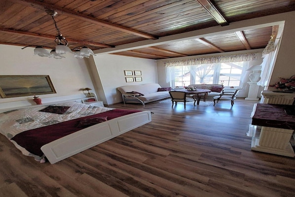A spacious room with a wooden ceiling, a bed, a sofa, a dining table with chairs, and a window with a view.