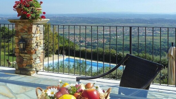 A balcony with a stone pillar, a wicker chair, and a table with fruit and flowers. - Estate in Monte Serra with Pool & Olive Trees (Pisa)