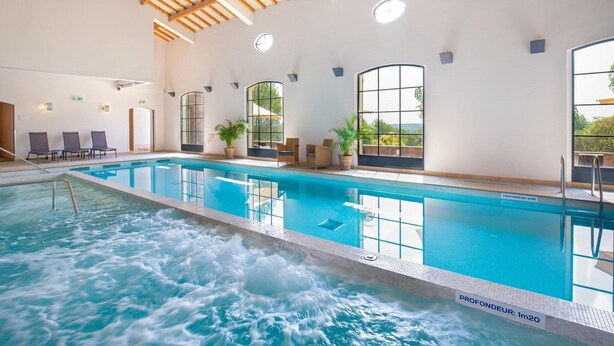 Une piscine intérieure dotée d’un espace jacuzzi, de poutres en bois au plafond et de grandes fenêtres.
