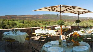 Un espace de restauration en plein air, avec des tables disposées pour deux personnes, entouré de murs en pierre et offrant une vue panoramique sur un paysage pittoresque composé de collines et d’arbres.