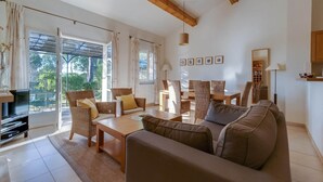 Un salon spacieux comprenant un canapé, des fauteuils, une table basse et un téléviseur. De grandes fenêtres sont agrémentées de rideaux, et on y trouve également une bibliothèque et une lampe.