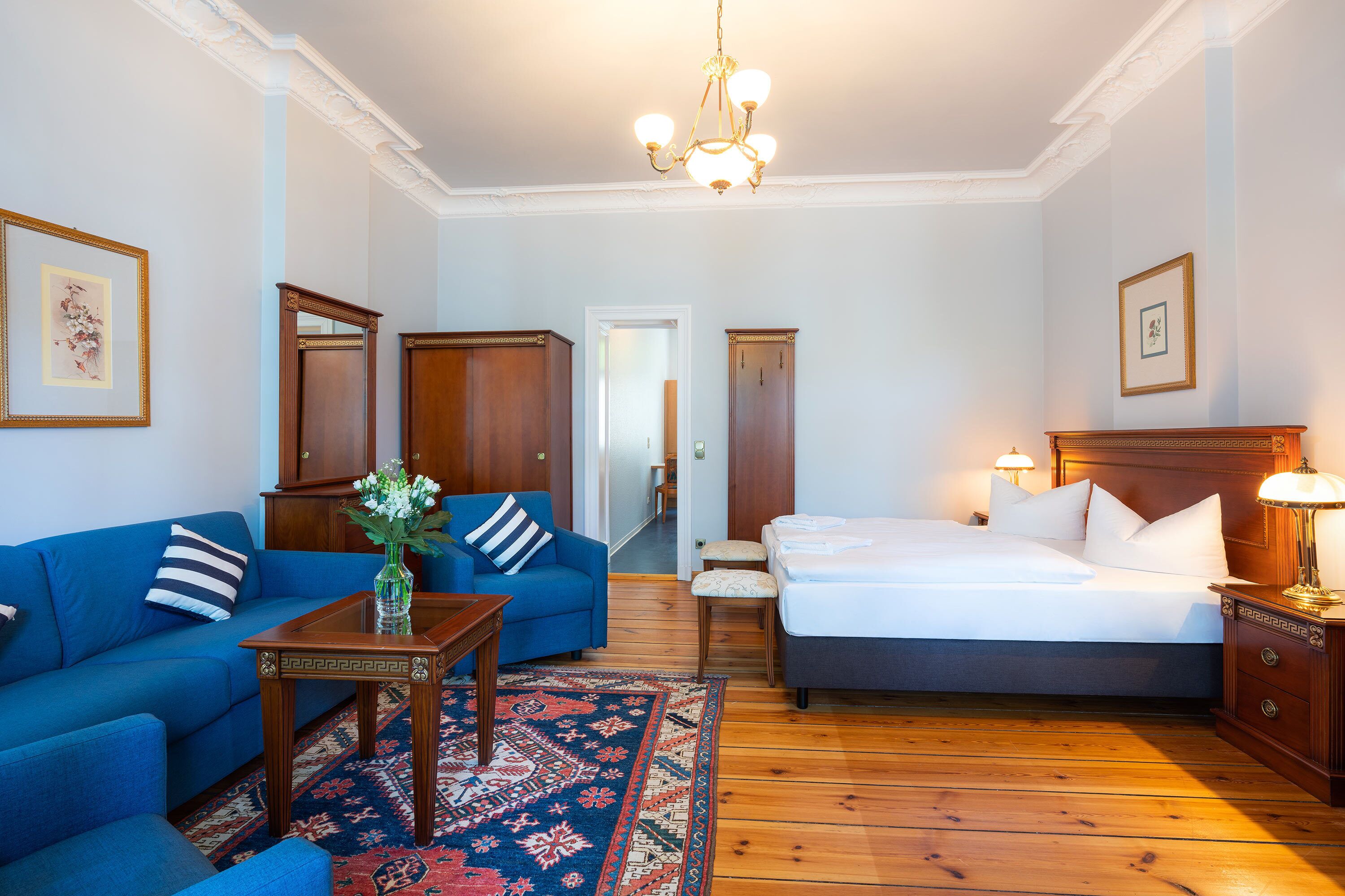 A hotel room with a blue sofa, a wooden bed, a nightstand with a lamp, a wooden floor, and a hallway leading to another room.