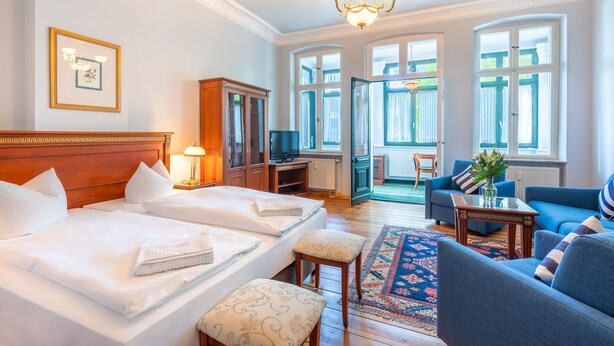 A hotel room with two beds, a sitting area with a sofa and armchairs, a television, and a large window with curtains. - SEETELHOTEL Villa Waldesruh (Heringsdorf)