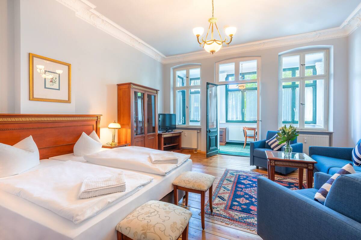 Habitación de hotel con dos camas, un área de descanso con sofá y sillones, un televisor y un ventanal con cortinas.