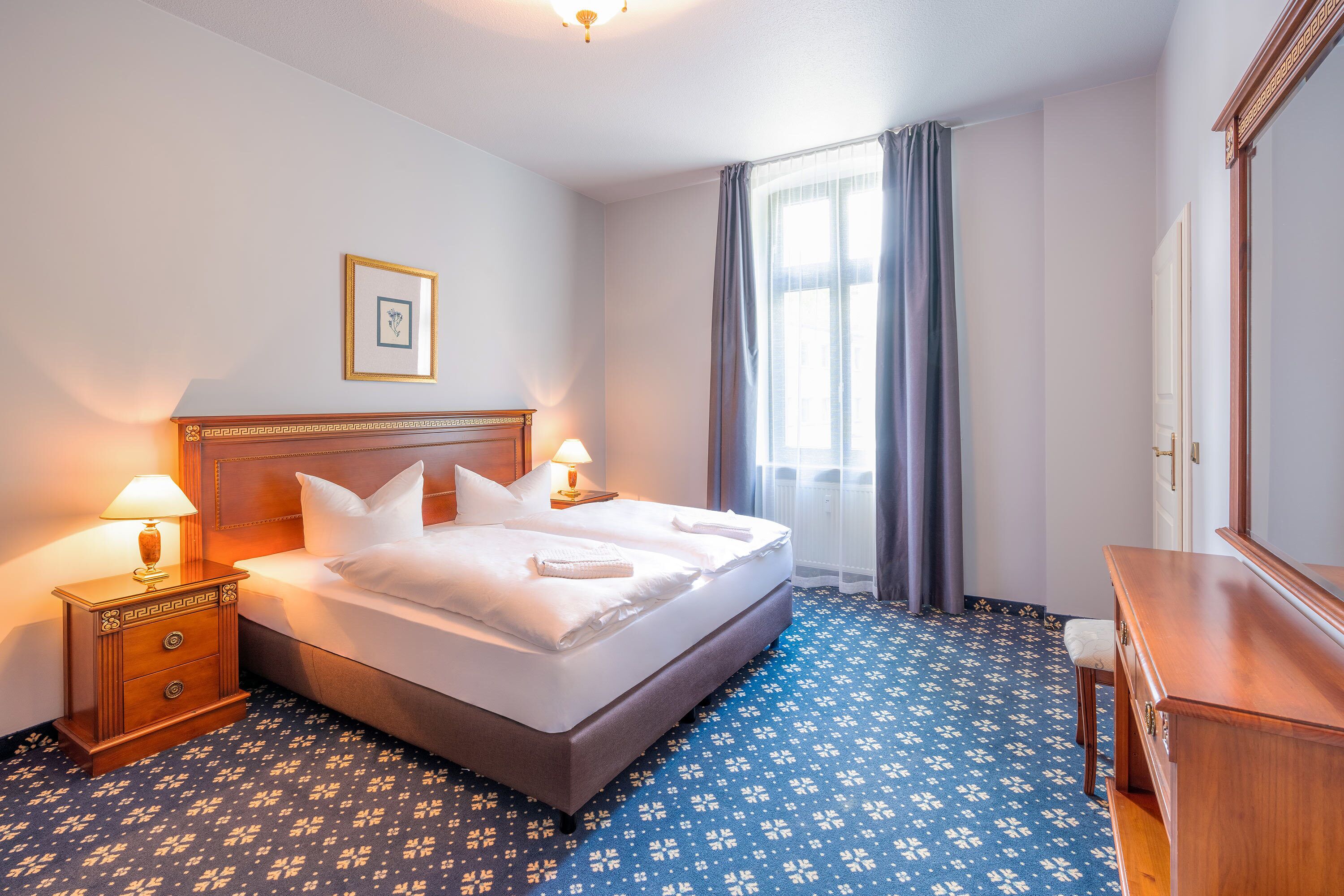 A hotel room with a bed, nightstand, dresser, and a window with curtains.