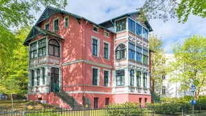 A multi-story pink building with a balcony, a staircase, and a fenced garden area. - SEETELHOTEL Villa Waldesruh (Heringsdorf)