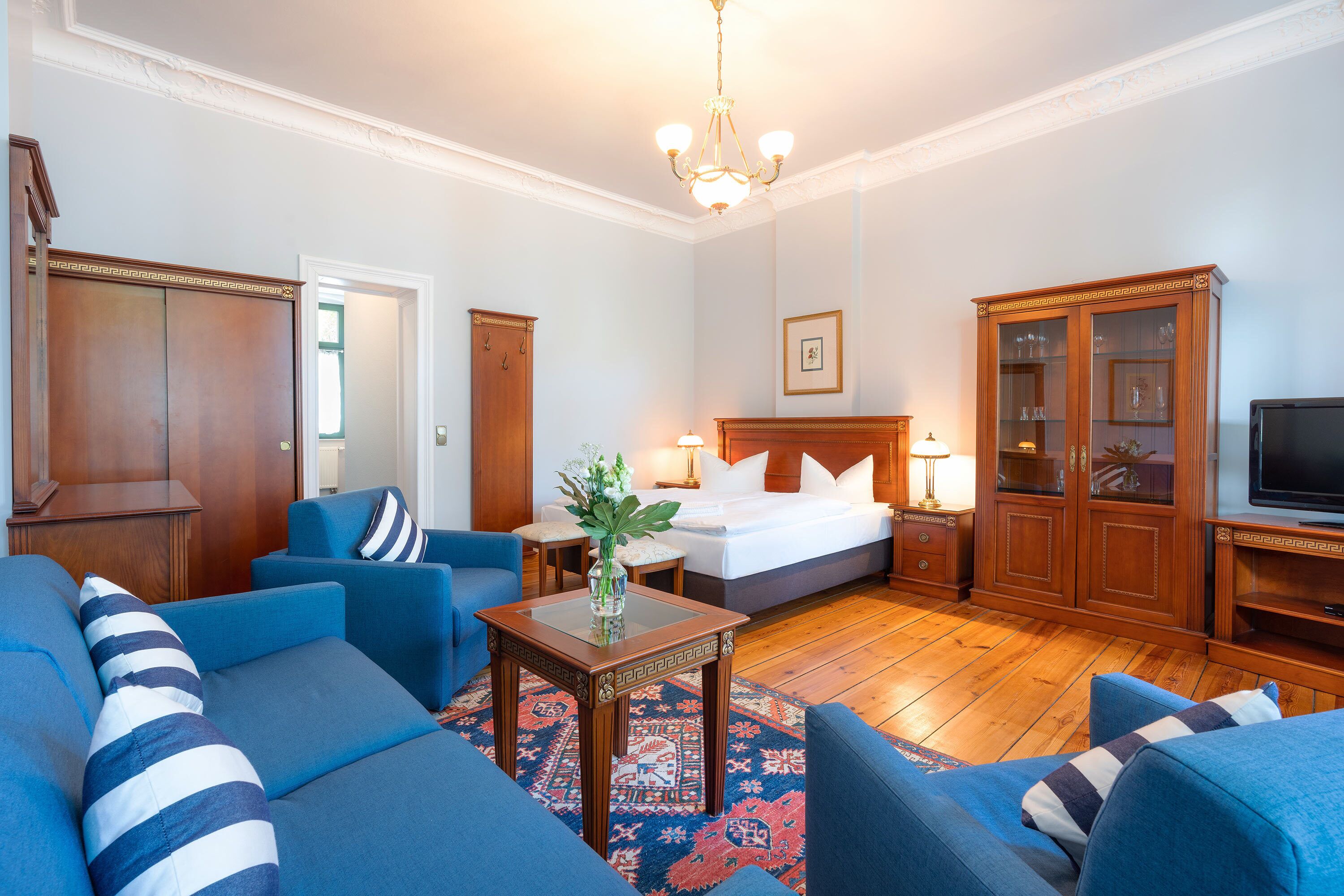 A hotel room with a bed, two blue sofas, a wooden cabinet, a television, and a chandelier.