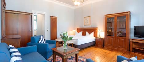 A hotel room with a bed, two blue sofas, a wooden cabinet, a television, and a chandelier.