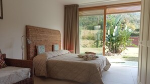 A bedroom with a large bed, a wicker chair, and a view of a garden through a sliding glass door.