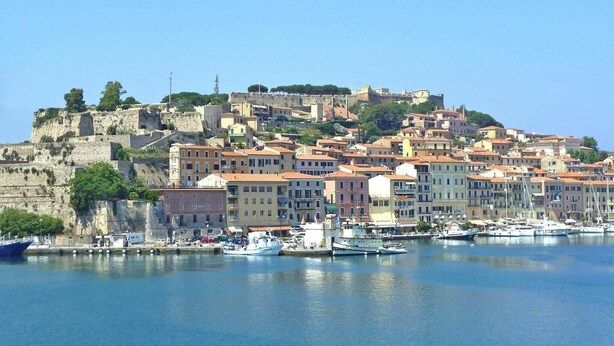 Un pueblo costero con edificios históricos, una colina fortificada y un puerto deportivo con barcos.