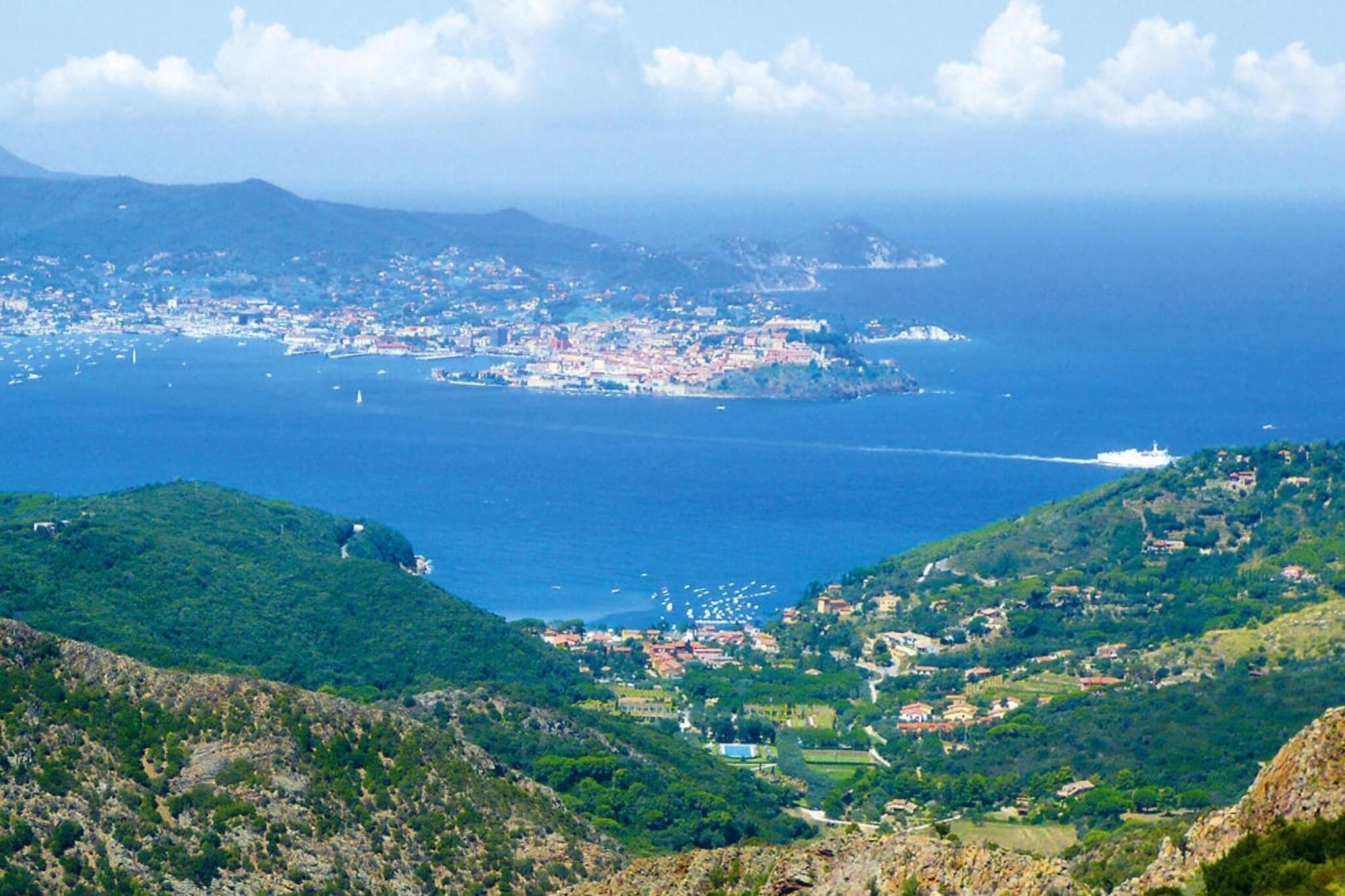 A coastal town with a mix of residential and commercial buildings, a prominent hill with a road leading up to it, and a view of the sea with boats.