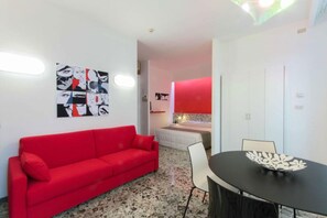 A modern hotel room with a red sofa, a dining table with chairs, a bed with a red headboard, and a wall with a pop art print.