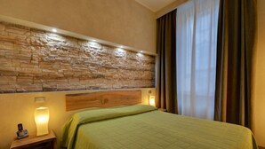 Une chambre d’hôtel avec un mur en pierre, une tête de lit en bois, une couvre-lit verte, une table de chevet avec une lampe et une fenêtre avec des rideaux.