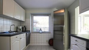 A compact kitchen with white cabinets, a stainless steel refrigerator, a coffee maker, and a window with blinds.