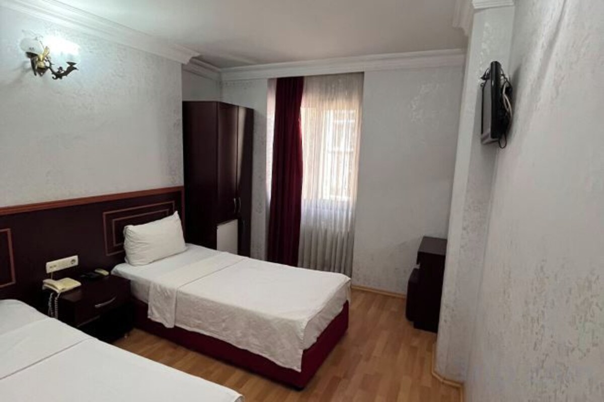 A hotel room with two beds, a wardrobe, a bedside table with a lamp, and a window with curtains.