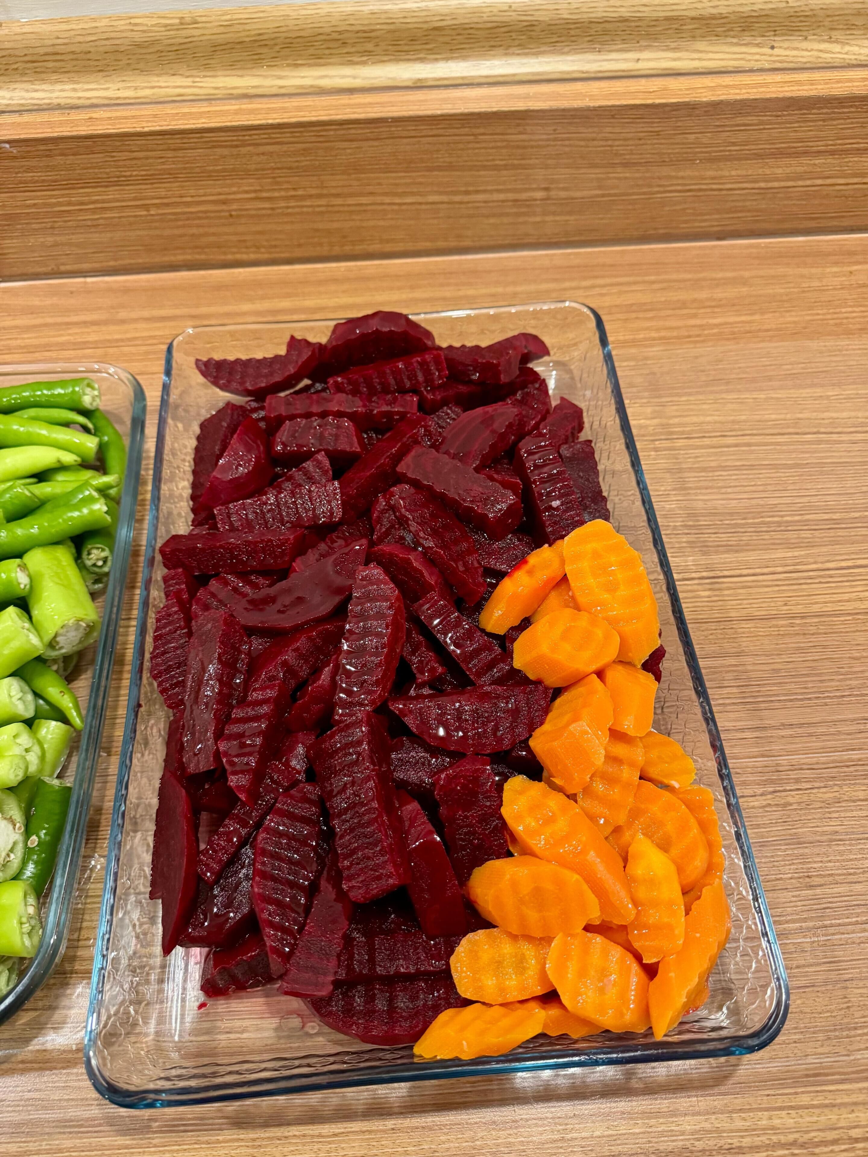 A glass dish with sliced beets, carrots, and green peppers on a wooden surface.