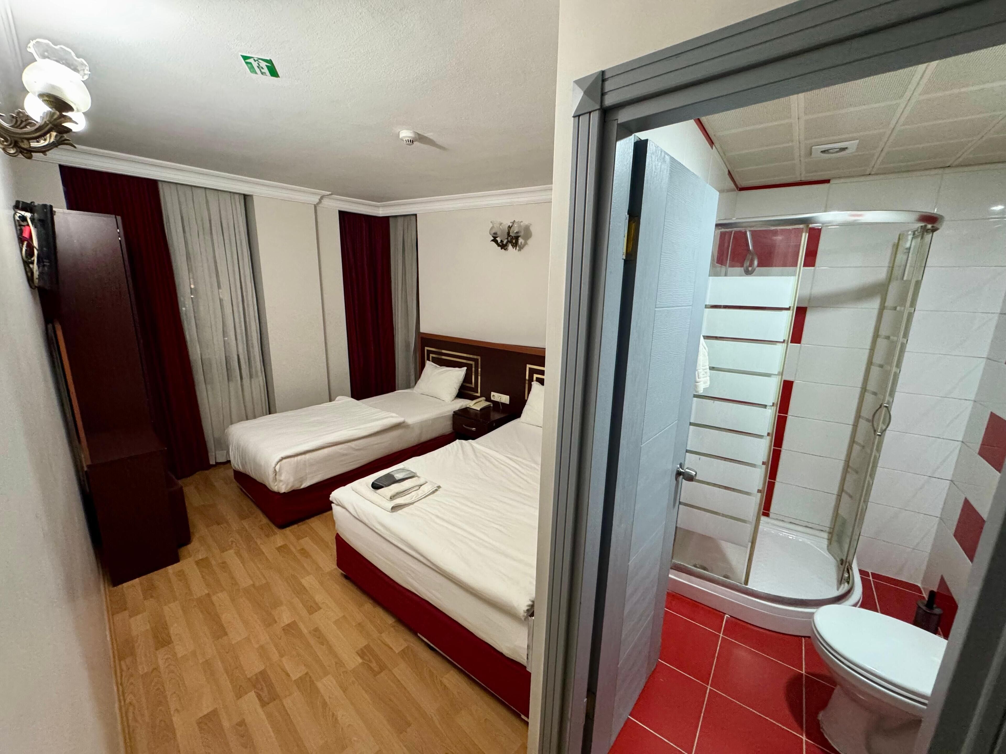 A hotel room with two single beds, a wooden floor, a red curtain, a white wall, a small wooden nightstand, and a bathroom with a red-tiled floor and white fixtures.