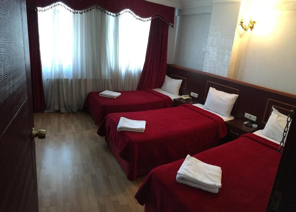 A hotel room with three beds, each with a red bedspread and white pillows. There is a wooden door on the left, a nightstand with a phone, and a wall-mounted light fixture.
