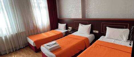 A hotel room with three beds, each with an orange bedspread, white pillows, and a wooden headboard. There is a window with sheer curtains and a red curtain partially drawn.