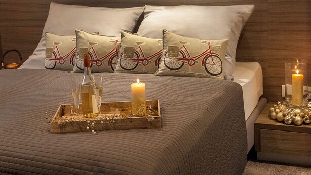 A bedroom with a wooden headboard, a quilted bedspread, and decorative pillows with a bicycle design. A tray with candles and a bottle is placed on the bed.