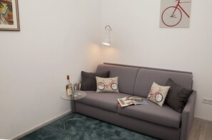 Ein modernes Wohnzimmer mit einem grauen Sofa, zwei Zierkissen, einem Glastisch mit Flasche und Gläsern sowie einer Wandleuchte.