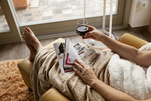 Eine Person entspannt auf einem Sofa, mit einem Buch und einem Glas Wein, in der Nähe eines Fensters mit Ausblick.