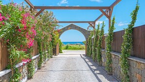 A pathway leading to a stone archway with a wooden pergola overhead, surrounded by flowering plants and a view of the sea. - Holiday home in Kiotari with a barbecue (Kiotari)