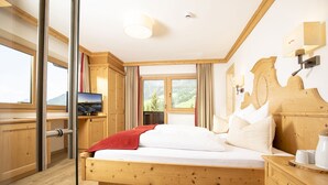 A wooden bed with a red blanket, a TV on a wooden cabinet, and a view of a mountain through large windows. - Alpbach Bergwald Apartments (Alpbach)