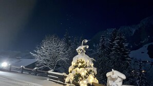 A snow-covered landscape with a Christmas tree adorned with lights, a wooden fence, and a mountain backdrop. - Alpbach Bergwald Apartments (Alpbach)