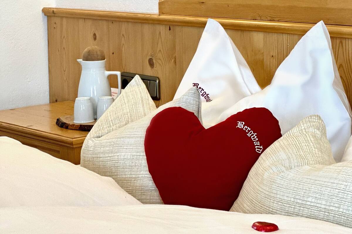 Una cama bien hecha con almohadas blancas y rojas, un cabecero de madera, una mesita de noche con una jarrón y tazas blancas, y una lámpara fijada a la pared.