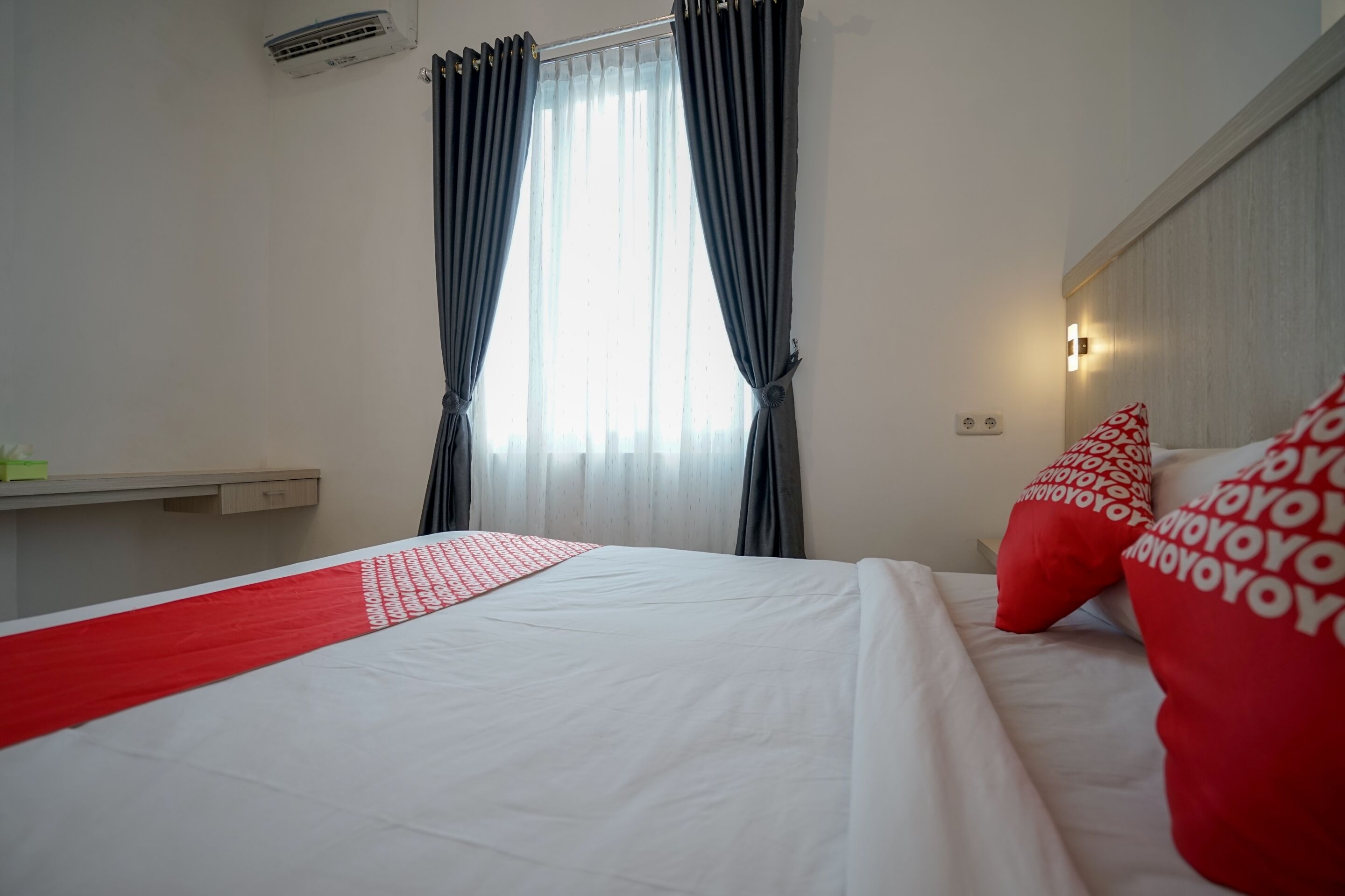 A hotel room with a bed, a window with curtains, a bedside table, and a wall-mounted light.
