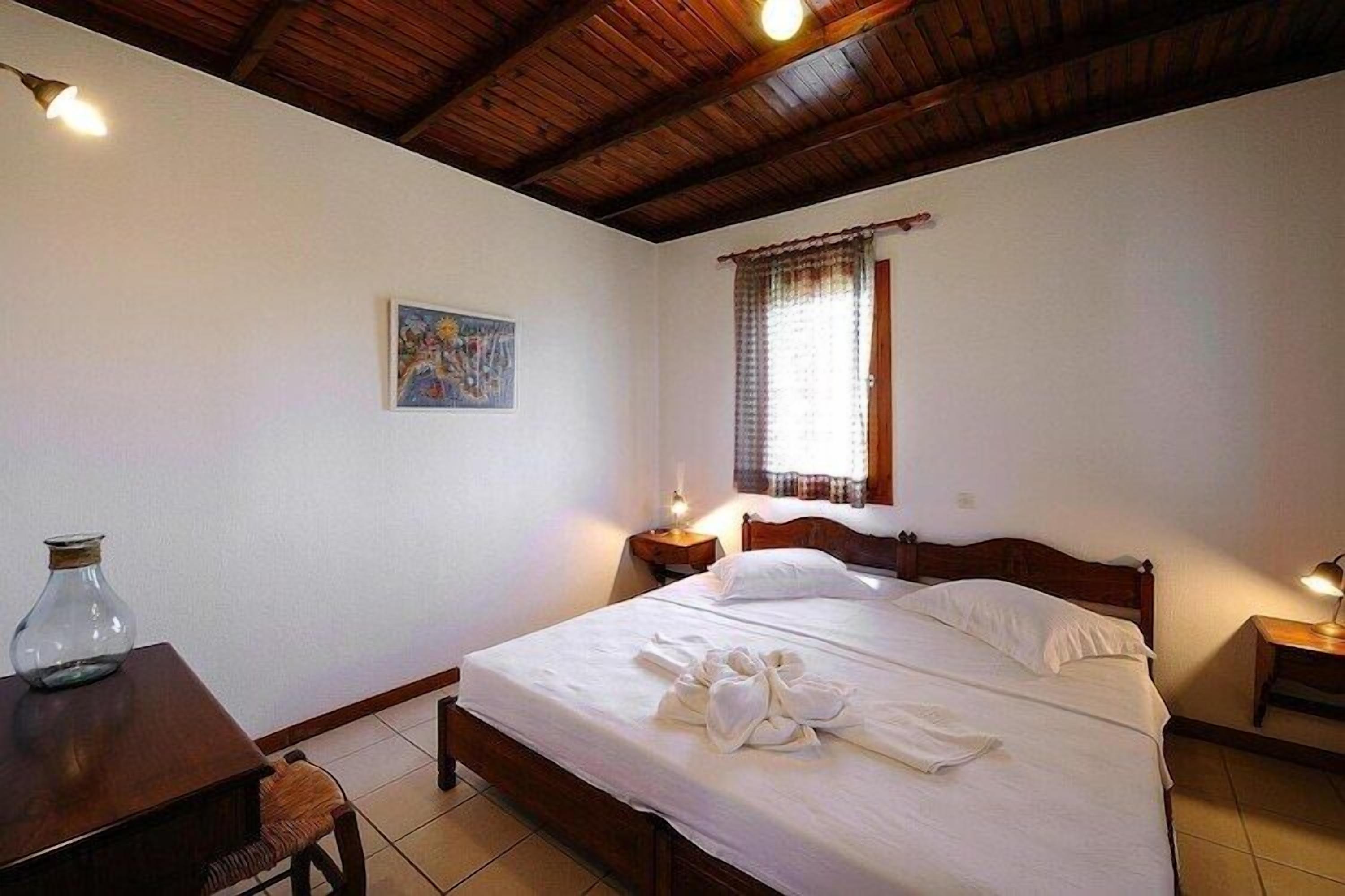 Un dormitorio con cama de madera, ropa de cama blanca, una mesita de noche con lámpara, una ventana con cortinas y un cuadro en la pared.