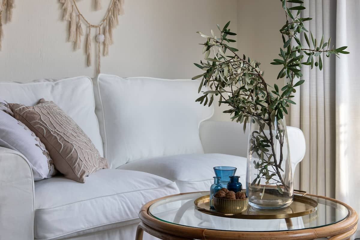Un sofá blanco con un colgador decorativo de macramé, una mesa de centro de vidrio con una planta y jarrones azules, y una ventana con cortinas translúcidas.