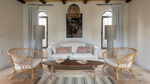 Una habitación con un sofá blanco, dos sillas de mimbre, una mesa de centro de madera y un ventanal con cortinas.