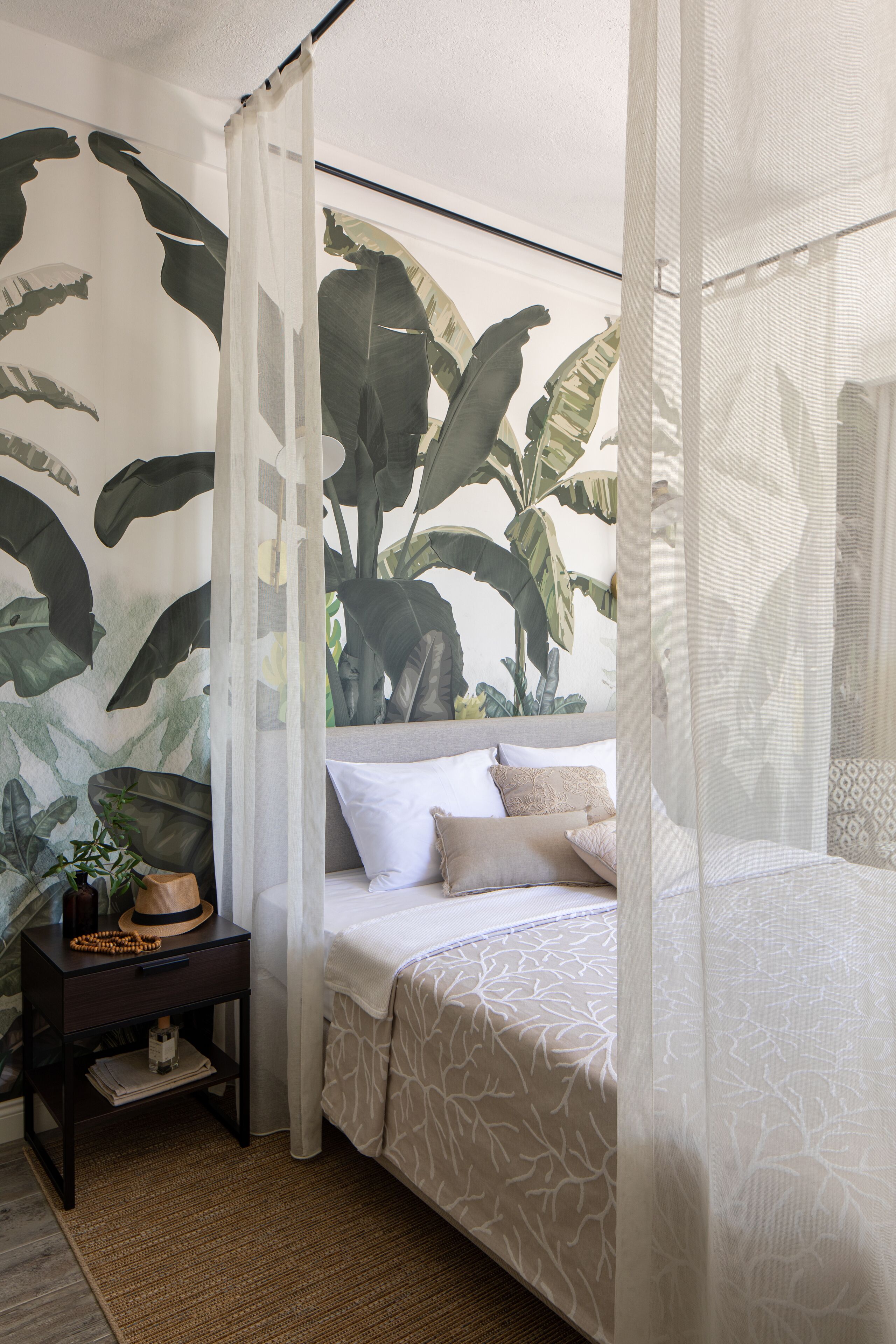 Un dormitorio con cama a dosel, mesita de noche con planta y un mural de hojas grandes.