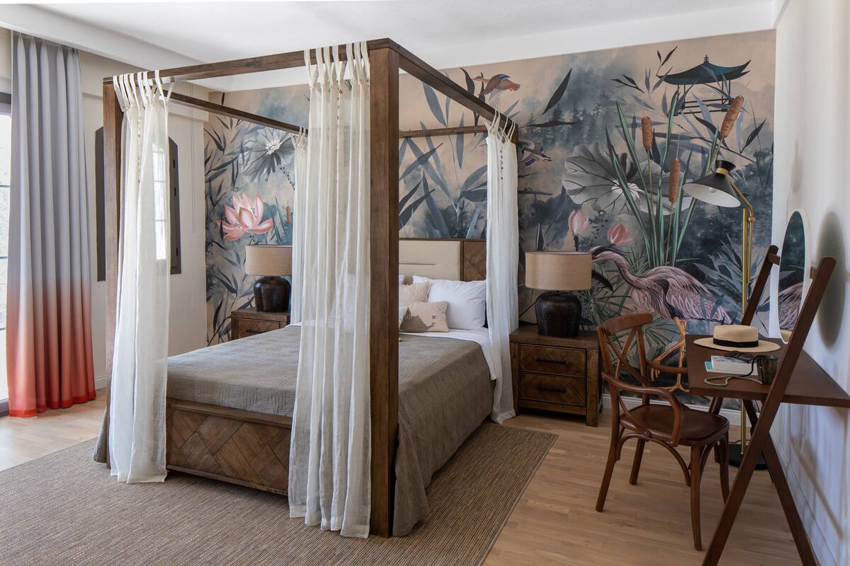 A four-poster canopy bed with a nightstand and chair, a mural with floral and bird designs, and a lamp on the nightstand.