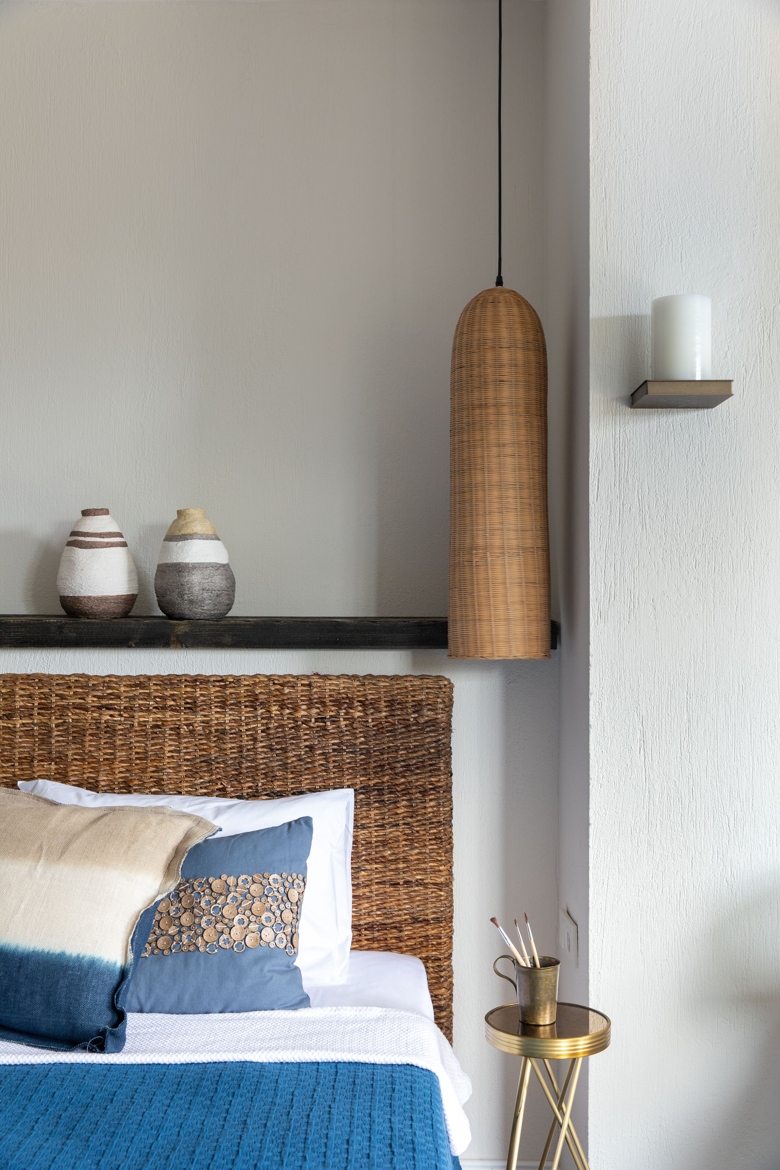 Un dormitorio ordenado con cama, almohadas, una mesita de noche, un estante en la pared con objetos decorativos y una lámpara colgante.