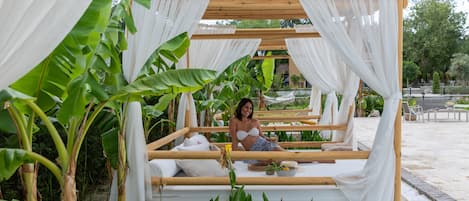 A wooden bed structure with white drapes, surrounded by lush greenery and banana plants.