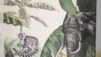 Quarto com um mural de um lêmure e um elefante, uma poltrona estampada e uma pequena mesa com uma planta e contas.